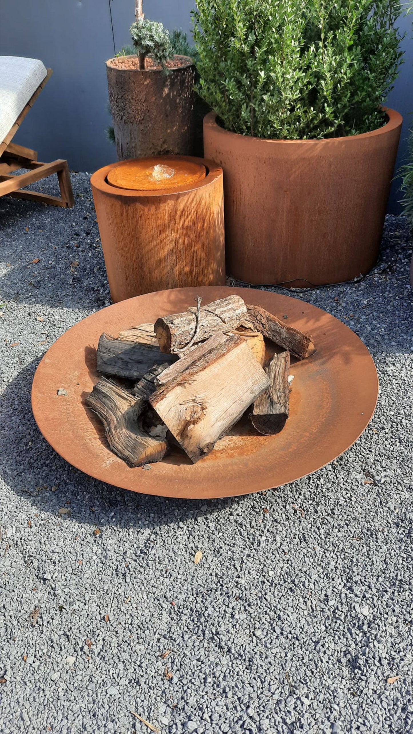 Ronde cortenstalen waterfontein naast vuurschaal en plantenbak